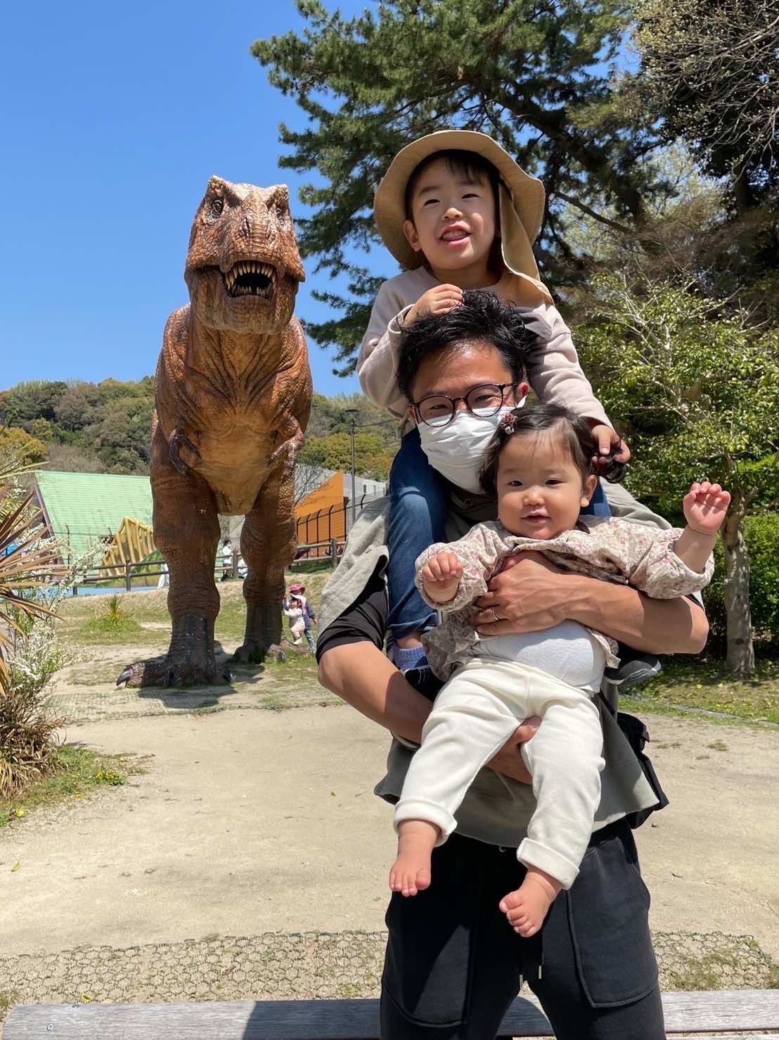 恐竜のモニュメントの前で子ども2人を抱っことおんぶしたパパ